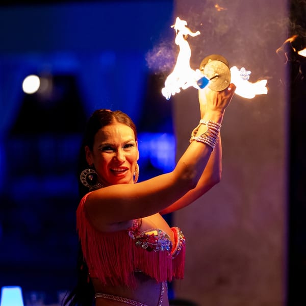 A smiling woman in a vibrant costume performs a fire dance with cymbals, illuminated by blue light.