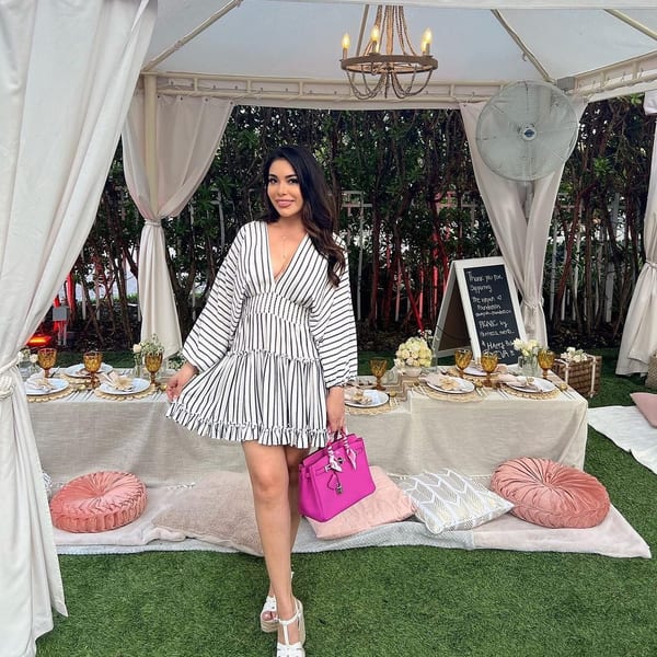 Woman in a striped dress holding a pink purse, standing by a picnic table under a white tent.