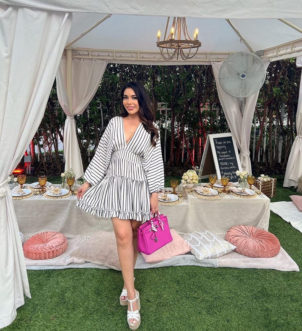 Woman in a striped dress holding a pink purse, standing by a picnic table under a white tent.