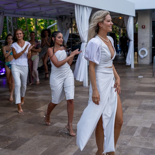 Models in white outfits and clear heels walk on a patio, with others clapping in the background.