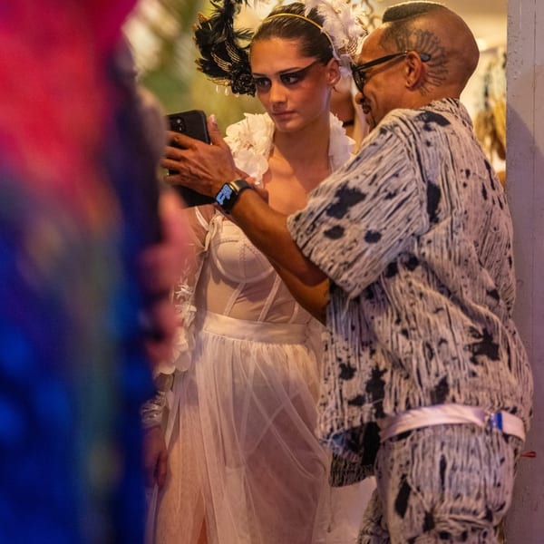 A model in a white feathered headpiece and sheer dress looks at a phone held by a man.