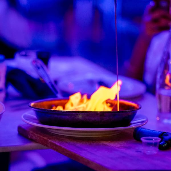 A gloved hand pours liquid from a jigger onto a flaming dish in a dark, blue-lit restaurant.