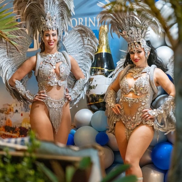 Two women in elaborate silver carnival costumes with feathers and sequins pose at a party.