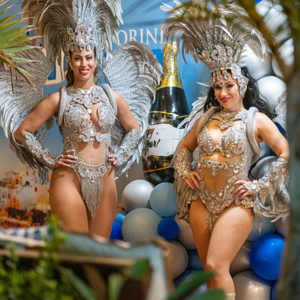 Two women in elaborate silver carnival costumes with feathers and sequins pose at a party.