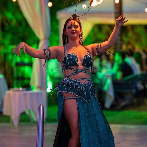Belly dancer in a teal costume with a flaming candelabra on her head, performing outdoors at night.